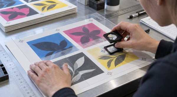 Technician using a magnifier to inspect registration on printed sheet samples.