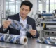 Editorial illustration of a Chinese businessman reviewing printed transfer-carrier material beside a matching finished sample in a workshop